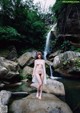 A naked woman standing on a rock in front of a waterfall.